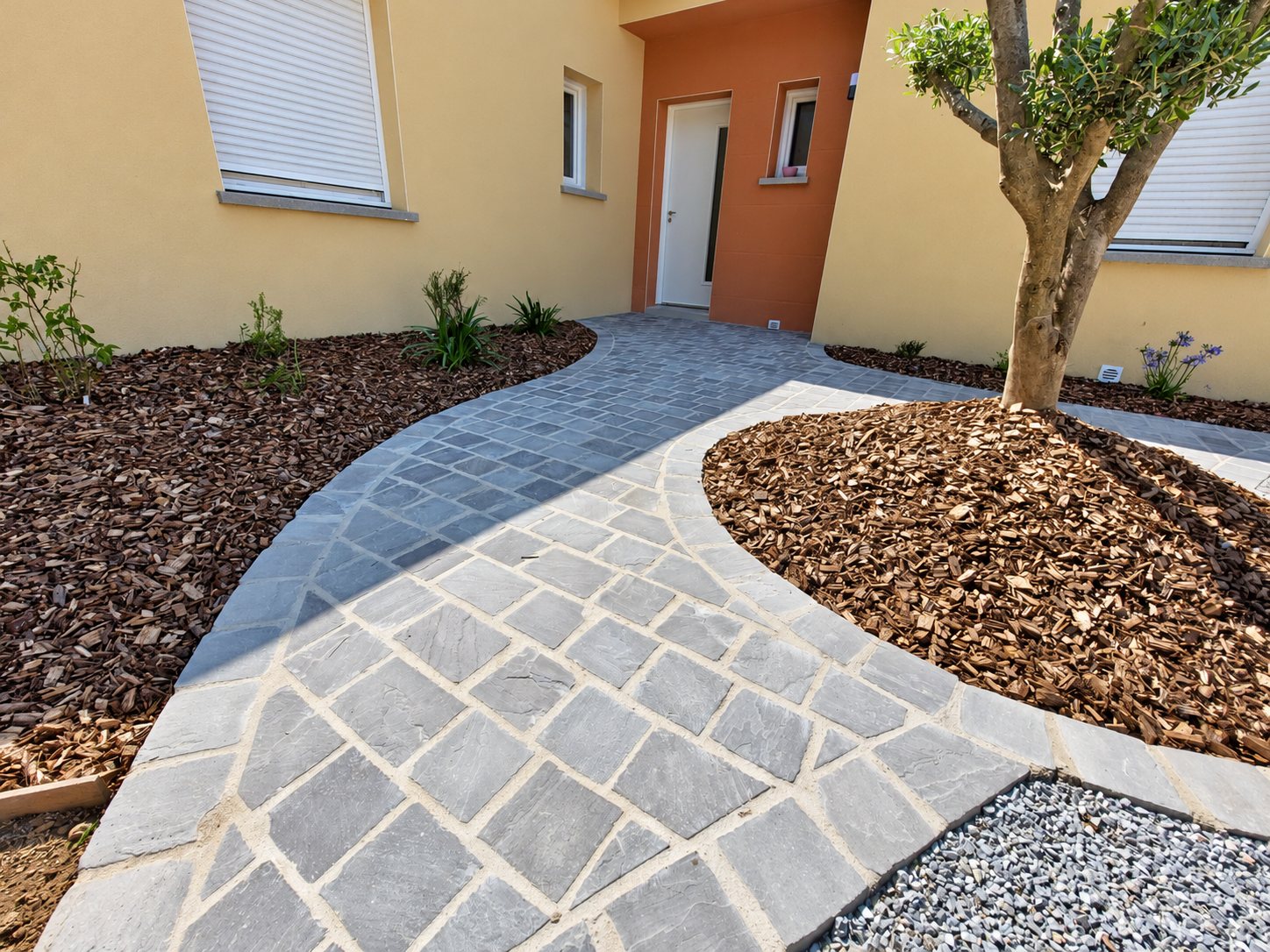 Une allée sinueuse en pavés de pierre grise mène à la porte d'une maison, entourée de massifs couverts de paillage et d'un olivier.