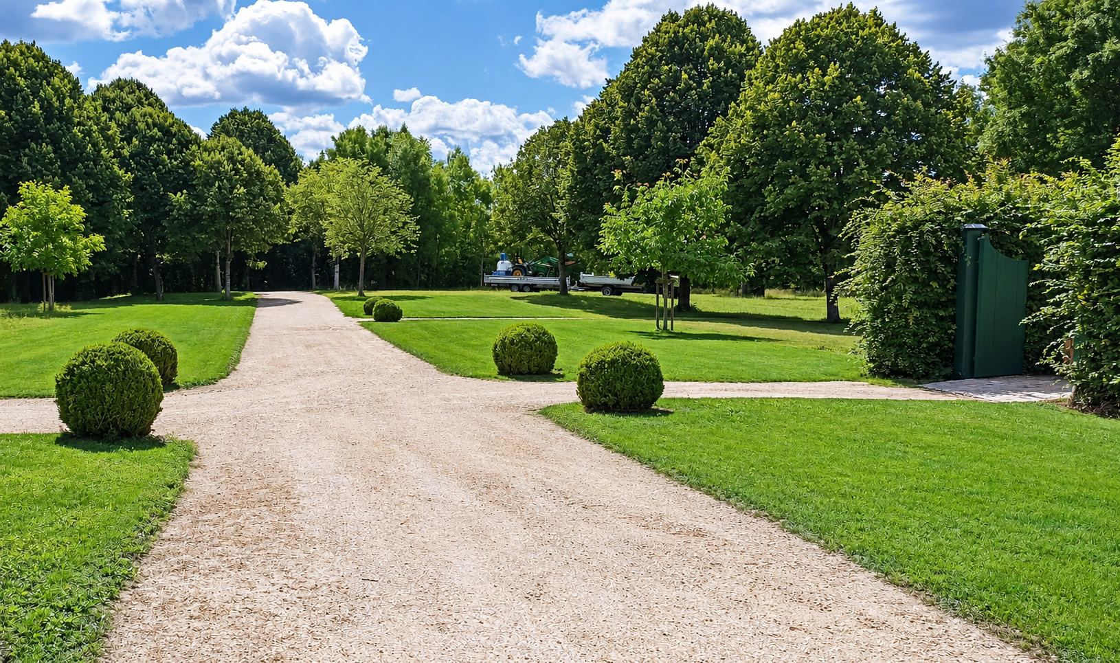 Une allée de gravier claire traverse une pelouse bien entretenue ornée de buis taillés en boule, entourée de grands arbres sous un ciel bleu.