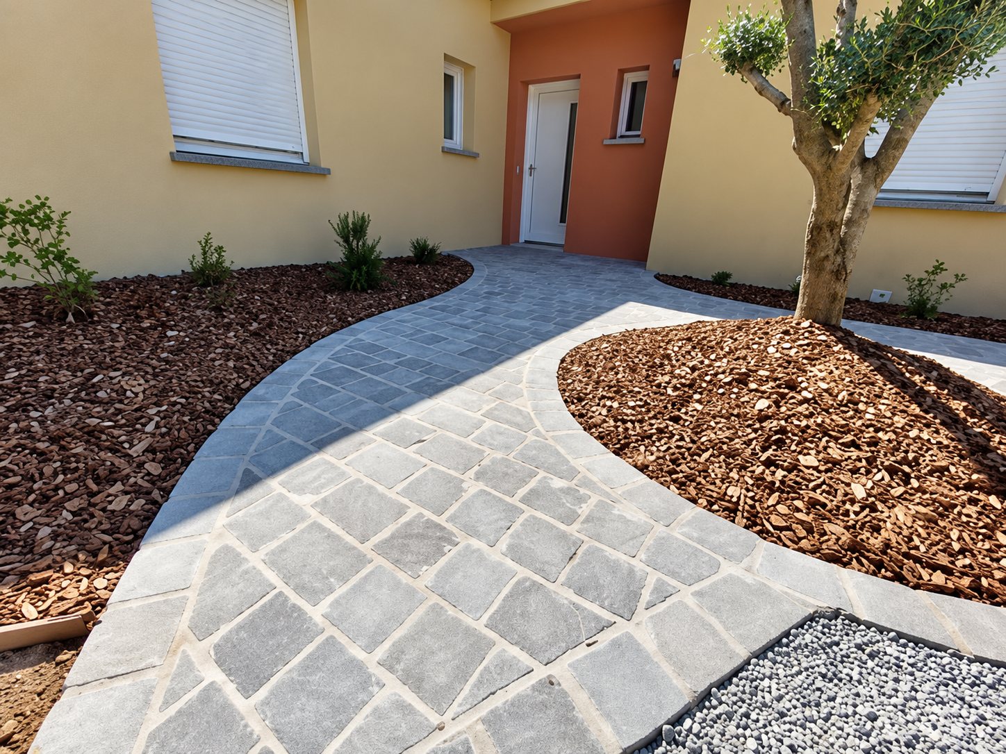 Une allée courbe en pavés de pierre grise mène à l'entrée d'une maison aux façades jaune et orangé, bordée de massifs couverts de paillage et d'un olivier.