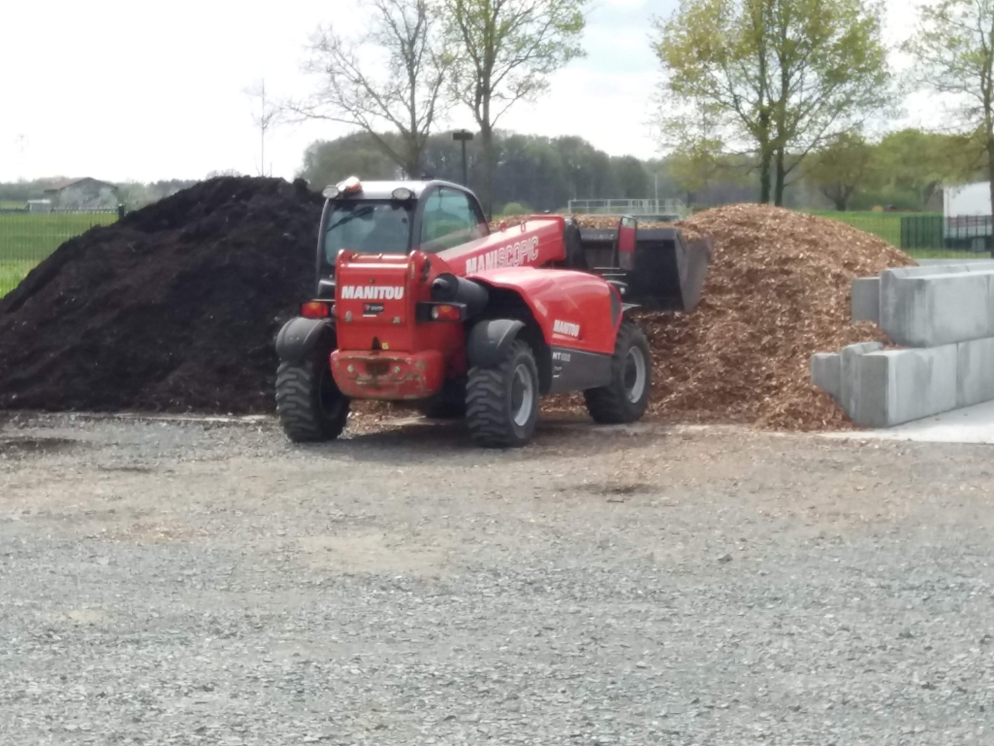 Un chariot télescopique Manitou rouge est garé sur du gravier devant un tas de terre noire et un tas de copeaux de bois, à côté de gros blocs en béton.