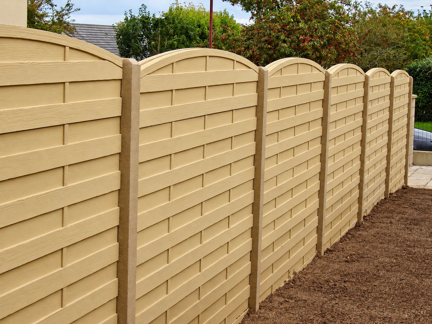 Une clôture beige composée de panneaux à lames entrelacées et au sommet arrondi, fixés entre des poteaux en béton le long d'une terre fraîchement remuée.