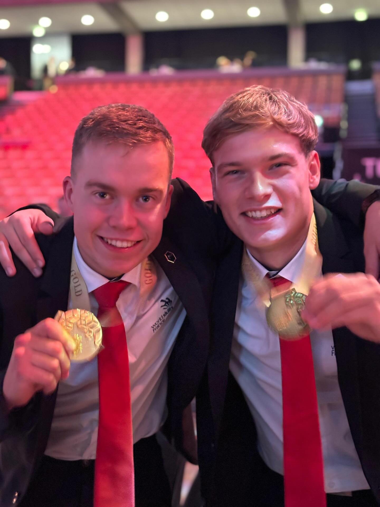 Deux jeunes hommes en costume sombre et cravate rouge posent en souriant et présentent chacun leur médaille d'or devant des gradins rouges.