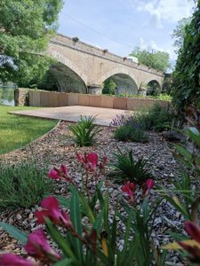 Un jardin aménagé avec des graminées, un paillage de copeaux et des fleurs roses au premier plan, une terrasse en bois et un vieux pont en pierre à arches en arrière-plan.