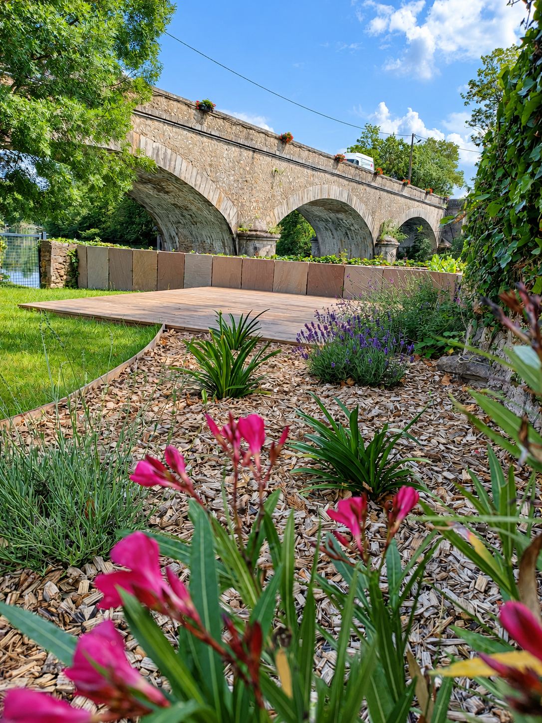 Un jardin aménagé avec une terrasse en bois, un paillage, des fleurs roses et de la lavande, au pied d'un grand pont en pierre.