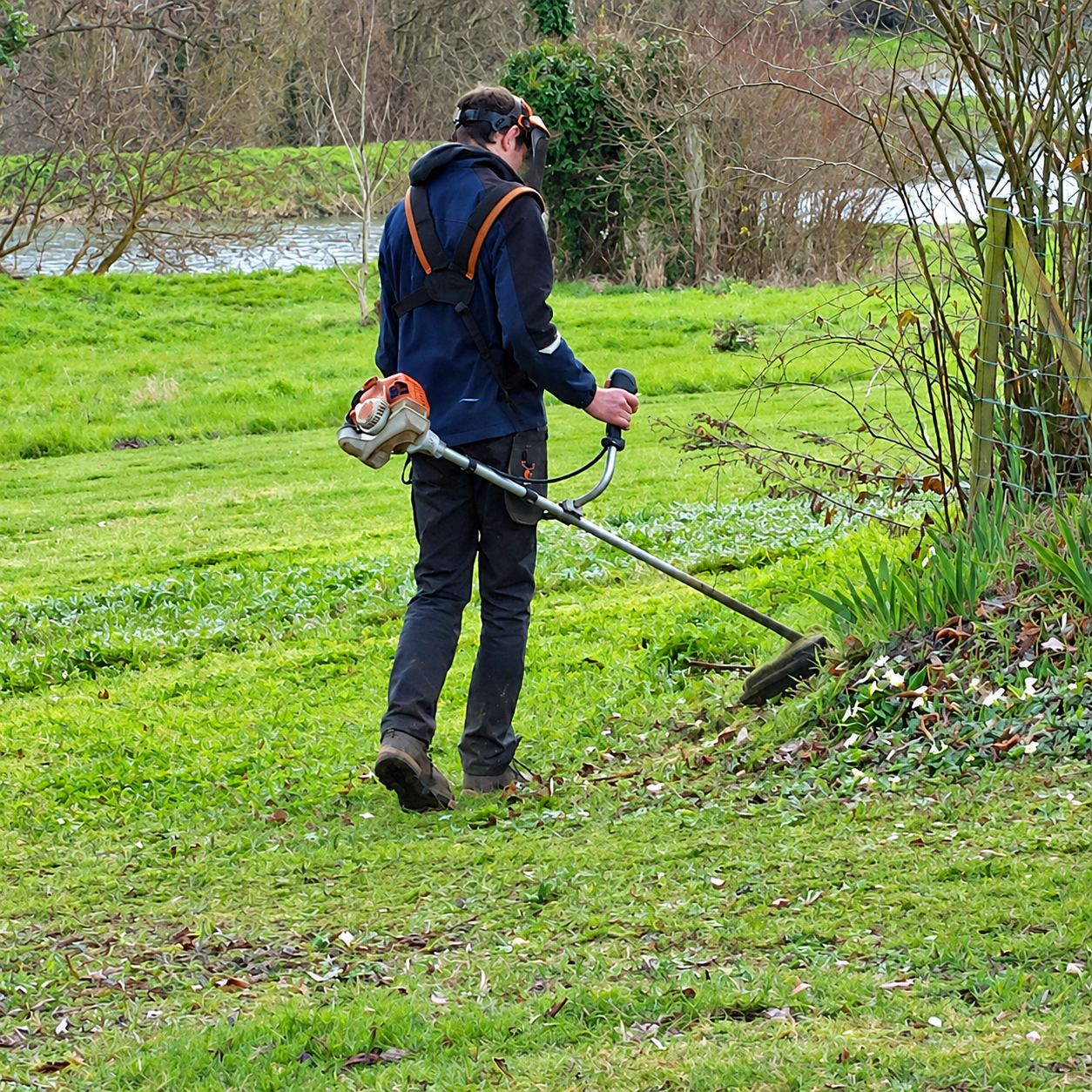 Un jardinier en veste bleue et casque coupe l'herbe avec une débroussailleuse sur une pelouse verte près d'une rivière.