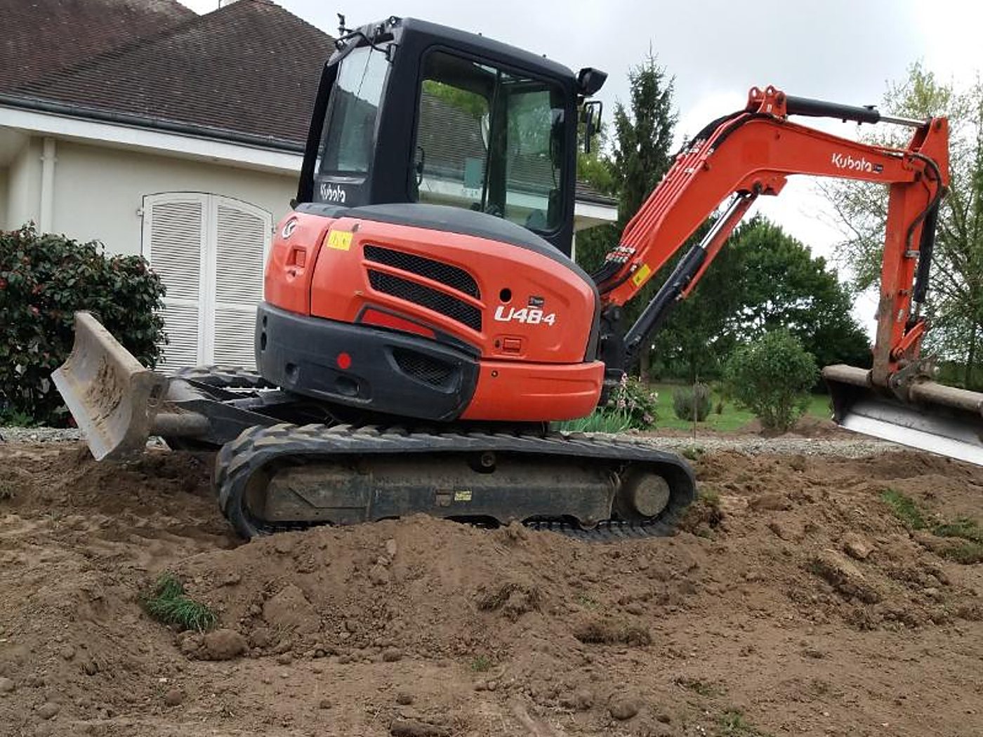 Une mini-pelle Kubota orange U48-4 sur chenilles travaille sur un terrain de terre fraîchement remuée devant une maison blanche.