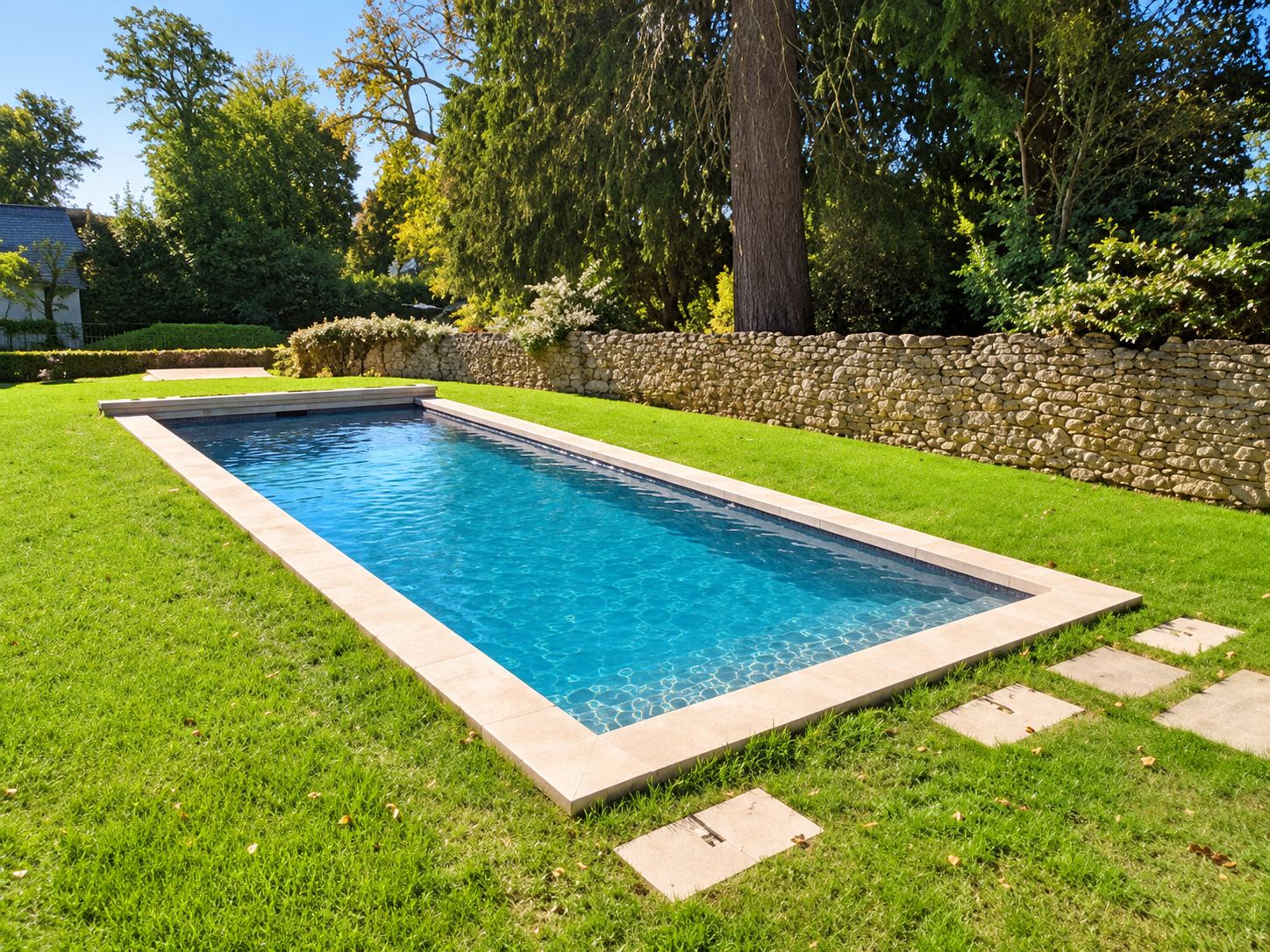 Une piscine rectangulaire à l'eau turquoise bordée de margelles en pierre claire, entourée de pelouse verte et d'un muret en pierres sèches.