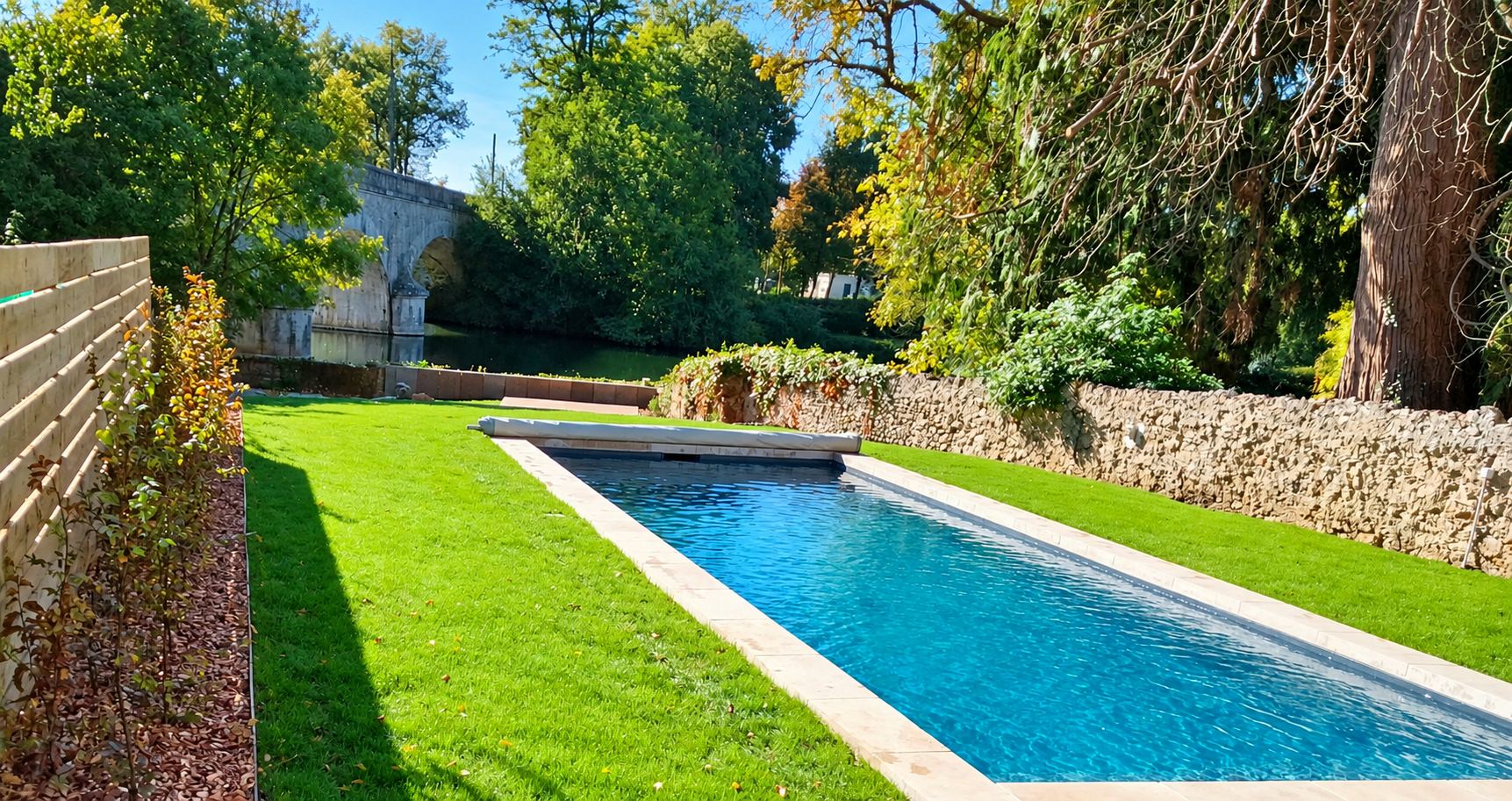 Une piscine étroite à l'eau turquoise, bordée de margelles claires, traverse un jardin de pelouse fraîche entre une clôture en bois et un mur en pierre, avec un pont ancien en arrière-plan.