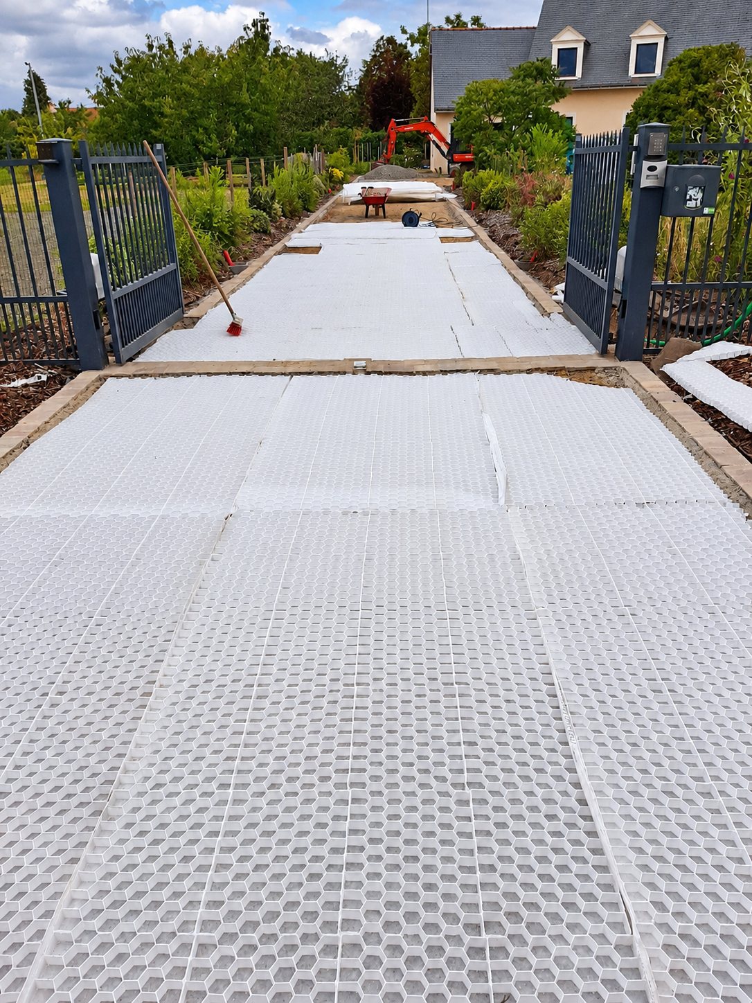 Chantier d'allée en cours avec des dalles alvéolaires blanches posées jusqu'à un portail gris, une mini-pelle orange et une brouette au fond.