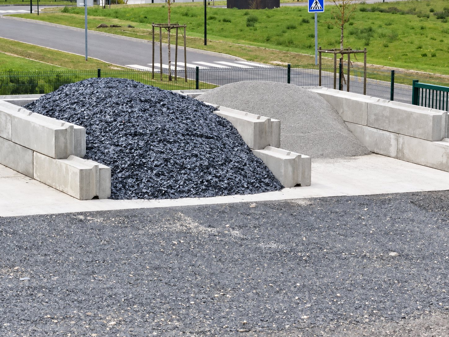 Deux tas de granulats, l'un en schiste sombre et l'autre en gravier clair, stockés dans des cases en blocs de béton au bord d'une route.