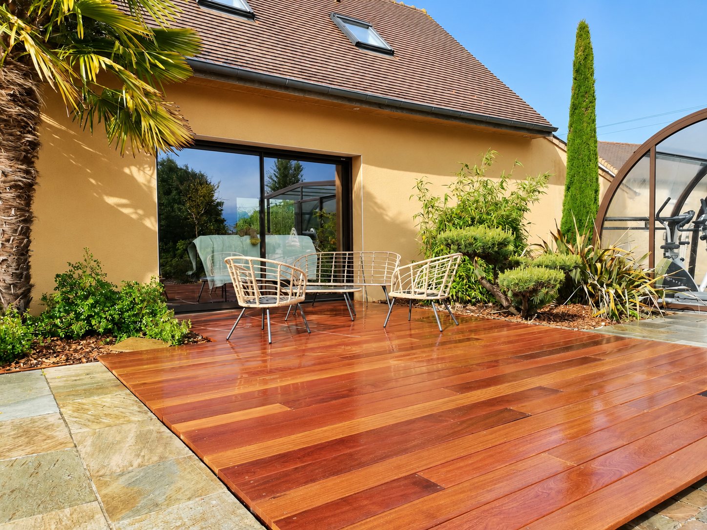 Une terrasse en bois exotique brillant, bordée de dalles en pierre, accueille trois fauteuils en rotin devant une maison ocre et un abri de piscine.