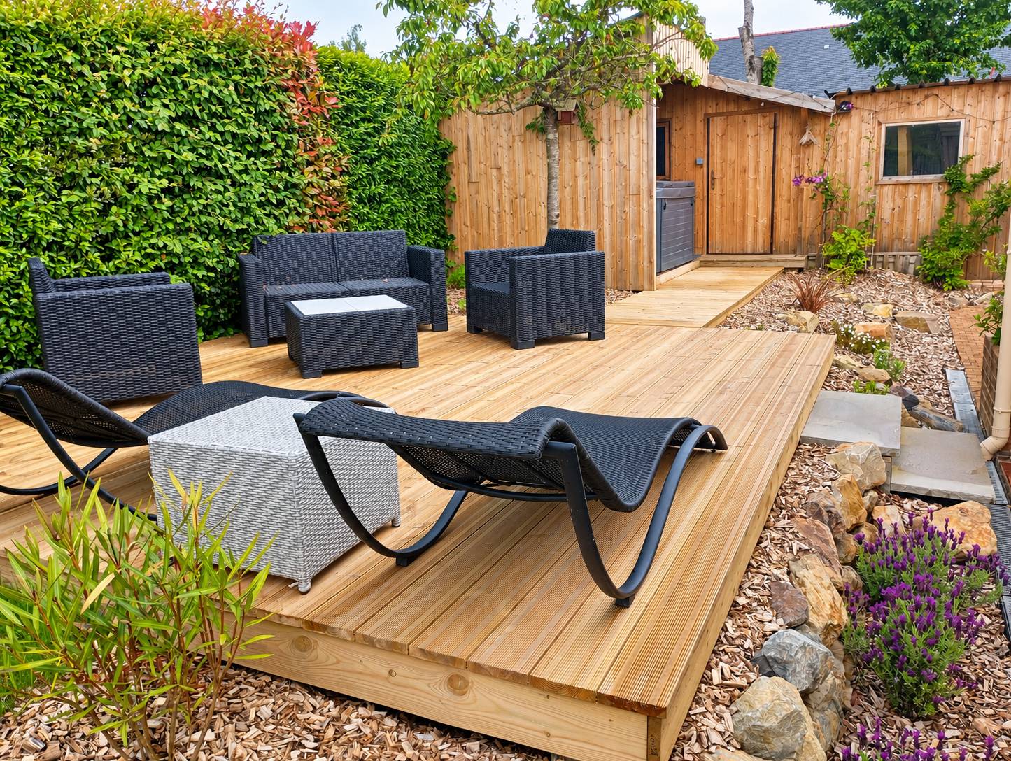 Une terrasse en bois dans un jardin, meublée d'un salon de jardin en résine tressée noire et de deux bains de soleil, entourée d'une haie et de massifs de pierres.