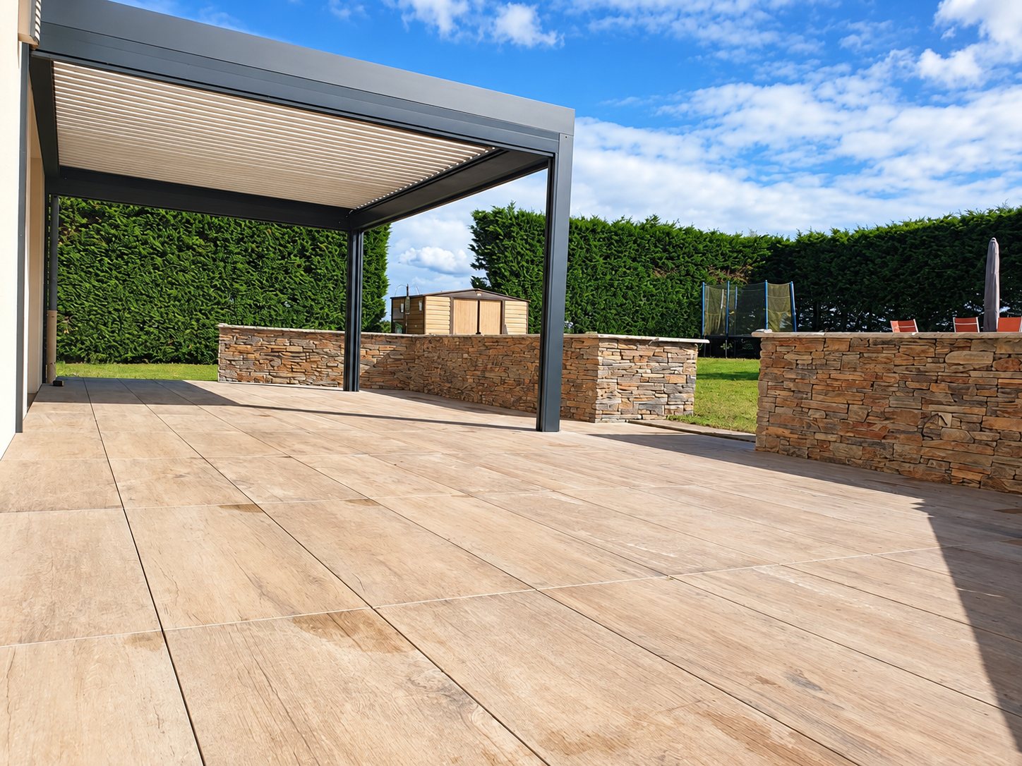 Terrasse en grands carreaux effet bois sous une pergola bioclimatique grise, bordée de murets en pierre et d'une haie dans un jardin.